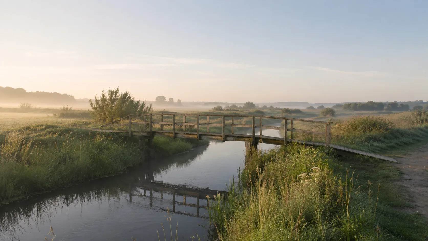 Lille slidt træbro over en stille å i dansk natur, blød morgendis og varmt morgenlys